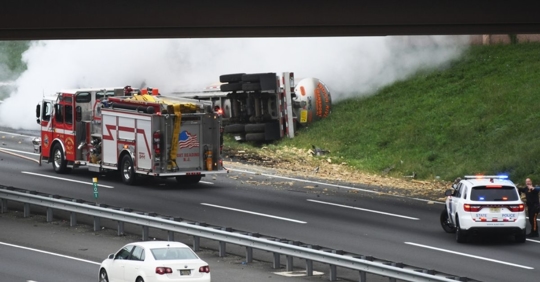 Photo of a truck accident under a highway overpass, smoke billowing, and a fire engine and police car on scene.