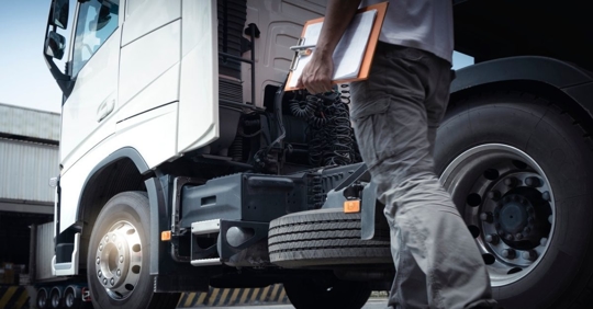 Individual in black jeans carrying a clip board walking alongside a white tractor trailer.